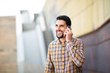 Dışarıda yürüyen, cep telefonuyla konuşan ve gülümseyen yakışıklı genç bir Arap portresi.