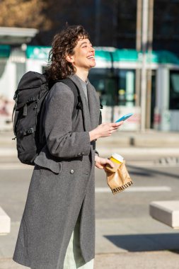 Cep telefonuyla yürüyen ve şehir dışına kahve fincanı götüren neşeli genç kadın portresi.