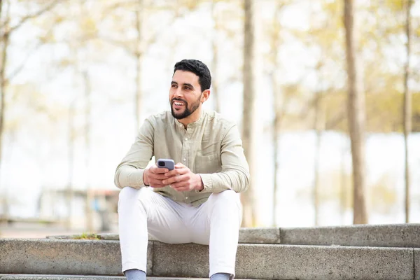 一个年轻的中东人坐在外面 带着手机朝外看 面带微笑的画像 — 图库照片