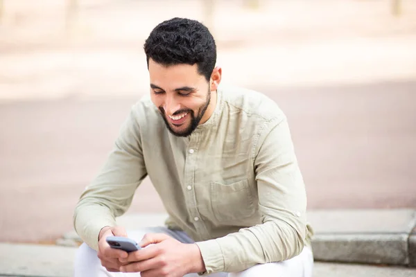 ハンサムな若いアラブ人の男の肖像画外に座っている彼の携帯電話を使用して笑顔 — ストック写真