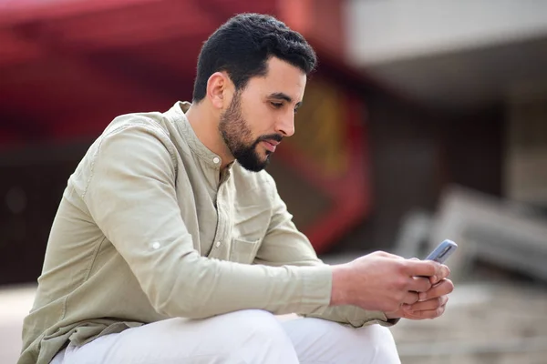 坐在外面看手机的阿拉伯年轻人的侧面肖像 — 图库照片