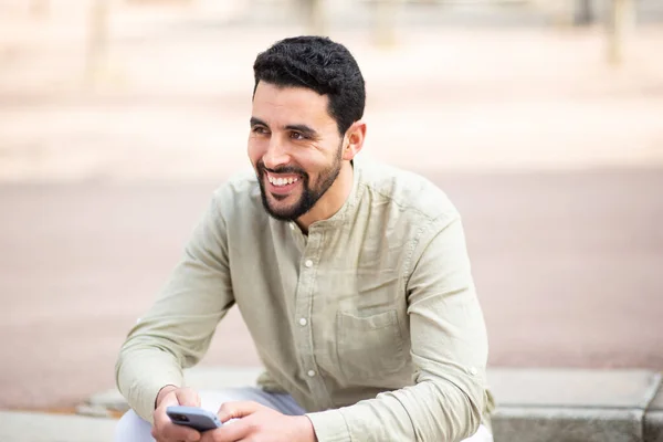 一个英俊的阿拉伯青年男子坐在外面 拿着手机 望着别处 笑着的画像 — 图库照片
