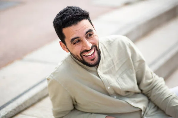 近照年轻英俊的中东男子坐在门外台阶上 望着远方笑着的画像 — 图库照片