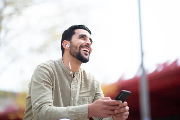手持耳机坐在外面听手机里的音乐 面带微笑的阿拉伯男子的画像 — 图库照片