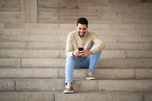 坐在台阶上看着手机笑着的阿拉伯青年男子的全长肖像 — 图库照片