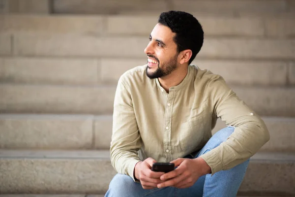 一个开朗的阿拉伯年轻人坐在台阶上 目不转睛地看着手机 微笑着的画像 — 图库照片