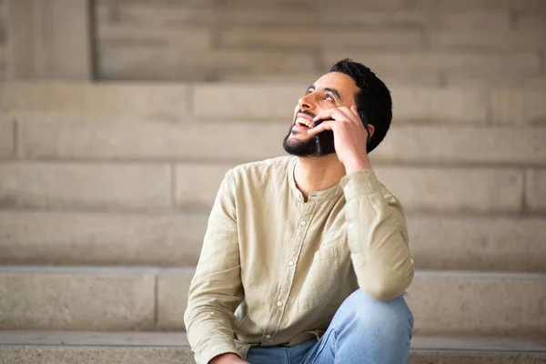 一个微笑的阿拉伯年轻人坐在台阶上用手机说话的画像 — 图库照片