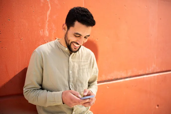 オレンジ色の壁に対する携帯電話でテキストメッセージを読む若い北アフリカの男の肖像画 — ストック写真