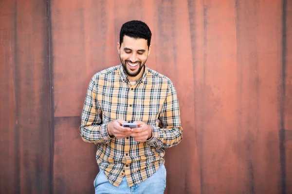 幸せな若いアラビア人の肖像画携帯電話を使用して茶色の壁に対して立って — ストック写真