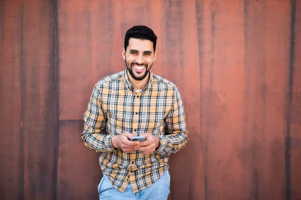若いアラビア人の笑顔の肖像画は茶色の壁に対して携帯電話で立って — ストック写真