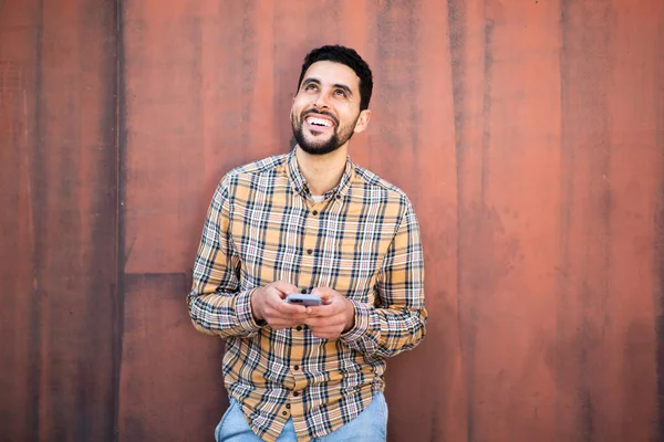 携帯電話で立っているハンサムな若いアラビア語の男の肖像画茶色の壁に対して離れて見て笑顔 — ストック写真