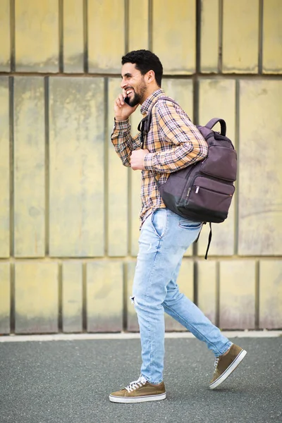 全长的侧面肖像英俊的年轻Arab的家伙走在外面与袋和手机交谈 — 图库照片