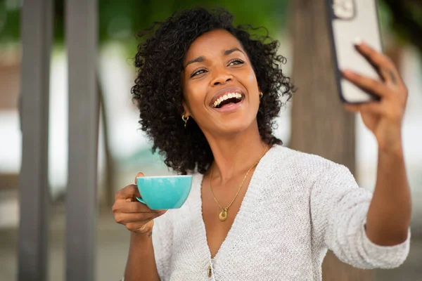 ポートレート若いアフリカ系アメリカ人女性保持カップのコーヒーと取りますセルフィーとともに携帯電話 — ストック写真
