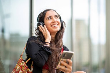 Cep telefonuyla oturup kulaklıkla müzik dinleyen gülümseyen kadın portresi