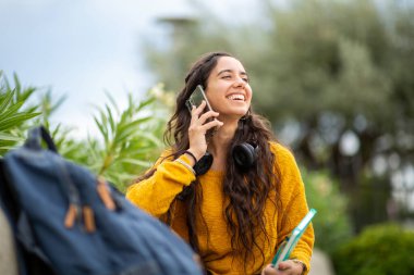 Çanta ve kitapla dışarıda otururken telefonla konuşan gülümseyen genç kadının portresi.