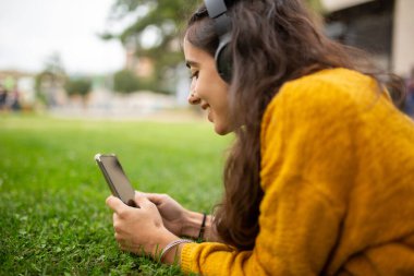 Kulaklık ve cep telefonuyla parkta müzik dinleyen genç bir kadının yan görüntüsü