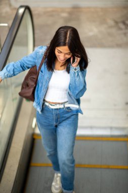 Yürüyen merdivende cep telefonuyla konuşan genç bir kadın portresi.