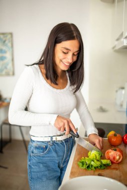 Mutfakta gülümseyen genç bir kadın. Bıçakla sebze doğruyor.