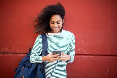 Portre gülümseyen Afrikalı Amerikalı genç bir kadın duvarda duruyor, cep telefonuna bakıyor.