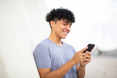 Beyaz duvarda cep telefonu kullanan gülümseyen genç adam portresi.