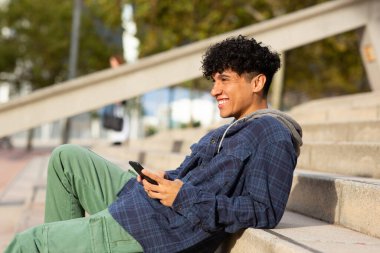 Yandan gülümseyen genç adam cep telefonuyla merdivenlerde oturuyor.