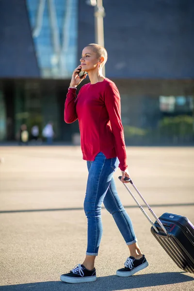 Full body profile portrait of travel woman walking with mobile phone and suitcase in city