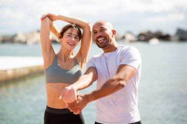 Zinde ve gülümseyen bir çift, deniz kenarında hafif bir esneme seansının tadını çıkarıyor.
