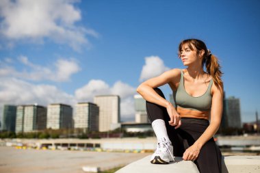 Güçlü ve kendine güveni tam bir kadın, açık mavi bir gökyüzünün altında, şehir siluetine karşı oturarak antrenmanına ara veriyor..