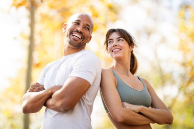 Kendine güvenen çift, sonbahar açık hava fitness oturumunda sırt sırta verip kollarını kavuşturur, dayanıklılık, ortaklık ve pozitiflik timsali olur.