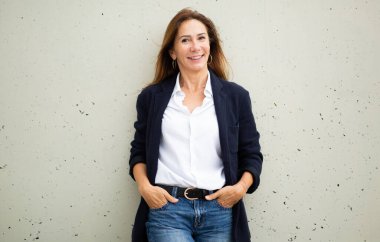 Kendine güvenen ve cana yakın bir kadın. Duvara yaslanmış, spor ceket, beyaz gömlek ve kot pantolon giymiş..