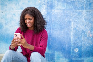 Kırmızı kazaklı kadın mavi duvarlı cep telefonuna bakıyor.