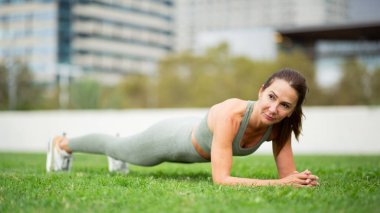 Portre formundaki kadın şehir parkında yeşil çimenler üzerinde bir planking egzersizi yapıyor.