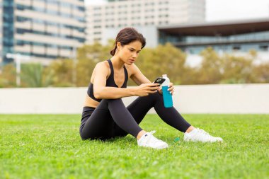 Şehir parkında çimlerde oturan, su içen ve spor yaptıktan sonra telefonu kullanan genç bir fitness kadını.
