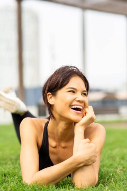 Neşeli genç Asyalı kadın çimenlerde uzanıyor, şehir parkında gülüyor.