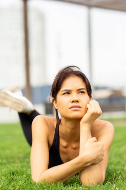 Genç Asyalı kadın çimlerin üzerinde uzanıyor, şehir parkında düşünceli bir ifadeyle uzaklara bakıyor.