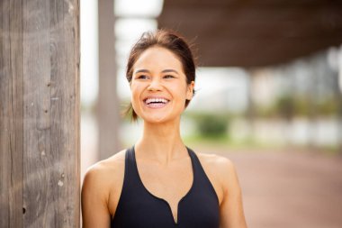 Atletik giyinmiş neşeli genç bir kadının yakın plan portresi. Dışarıda dururken gülümsüyor.