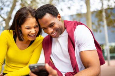 Çeşitli genç arkadaşlar bir akıllı telefona bakarken birlikte gülüyorlar. Neşeli bir Asyalı kadın ve gülümseyen bir zenci birlikte cep telefonuna bakıyor.