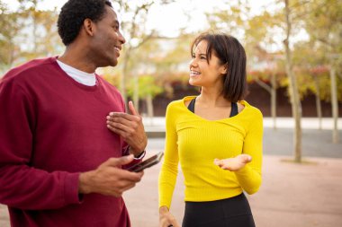 Genç çok kültürlü arkadaşlar dışarıda neşeli bir konuşma yapıyorlar. Siyahi bir adam ve Asyalı bir kadın yüz yüze, rahat bir şehir parkında gülümsüyor ve konuşuyorlar.
