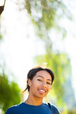 Açık havada, yumuşak güneş ışığında kameraya gülümseyen genç bir kadın.