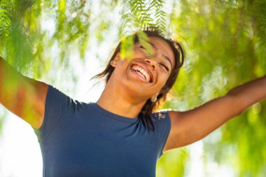 Yeşil dalların altında kollarını açmış gülen neşeli genç bir kadın portresi.