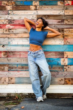 Elleri başının arkasında, duvarın yanında duran gülümseyen genç bir kadın.