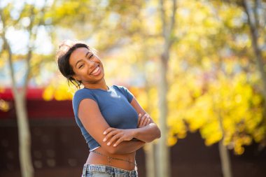 Genç bir kadın kollarını kavuşturmuş gülümsüyor, güneşli bir günde dışarıda dikiliyor.