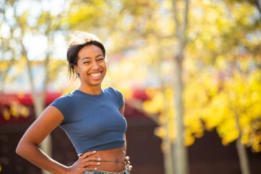 Güneşli bir sonbahar gününde, elleri kalçasında dışarıda duran gülümseyen genç bir kadın.