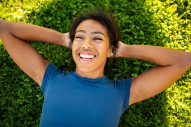 Genç bir kadın ellerini başının arkasına koyup yeşil çimlere uzanmış, gökyüzüne doğru gülümsüyor.