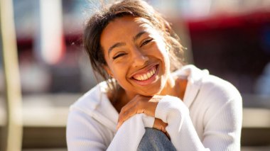 Gülümseyen genç bir kadın güneş ışığında dışarıda oturuyor.