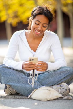Portre gülümseyen siyah bir kadın şehrin dışında oturuyor ve cep telefonuna bakıyor.