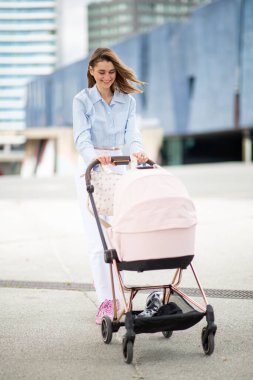 Tüm vücuduyla gülümseyen genç kadın bebek arabasını iterken modern bir şehir manzarasında yürüyor.