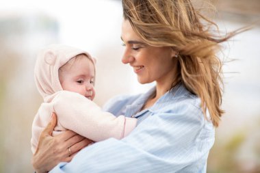 Gülümseyen anne, bebeğini kukuletalı kıyafetiyle kucaklıyor, açık havada hassas bir anı paylaşıyor.