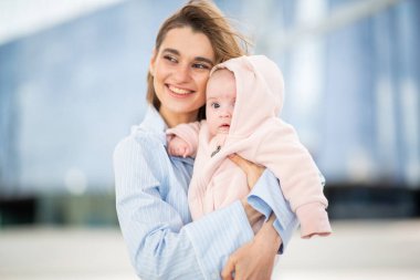 Gülümseyen anne, bebeğini sevgiyle kucağına alır. Yumuşak pembe bir tulum giyer.