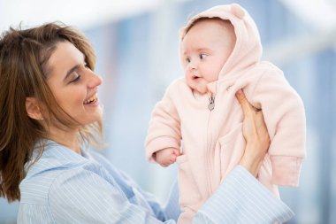 Gülümseyen anne bebeğini pembe kapüşonlu bir tulumla kucağına alıp açık havada neşeli bir anı paylaşıyor..